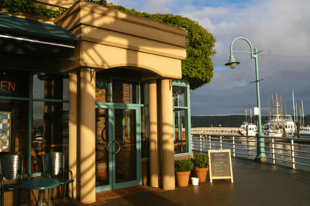 The exterior of a waterfront café with beige columns and teal-framed glass doors, surrounded by potted plants and a chalkboard sign. The golden sunset casts warm shadows across the boardwalk, with boats and lamp posts visible along the marina in the background.