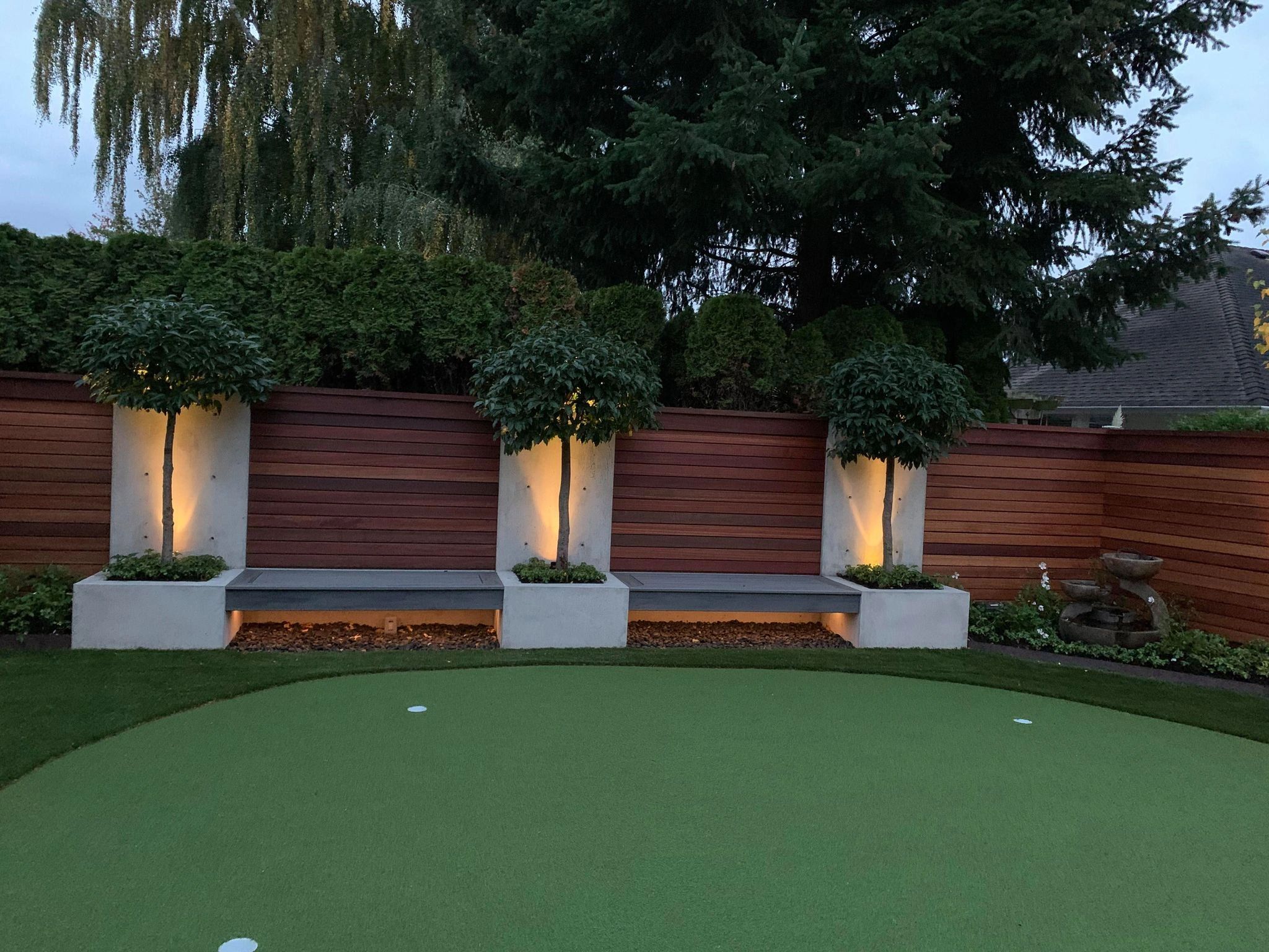Modern backyard landscape design in Port Moody with putting green, illuminated white planter boxes with topiaries, horizontal cedar fence, and mature trees