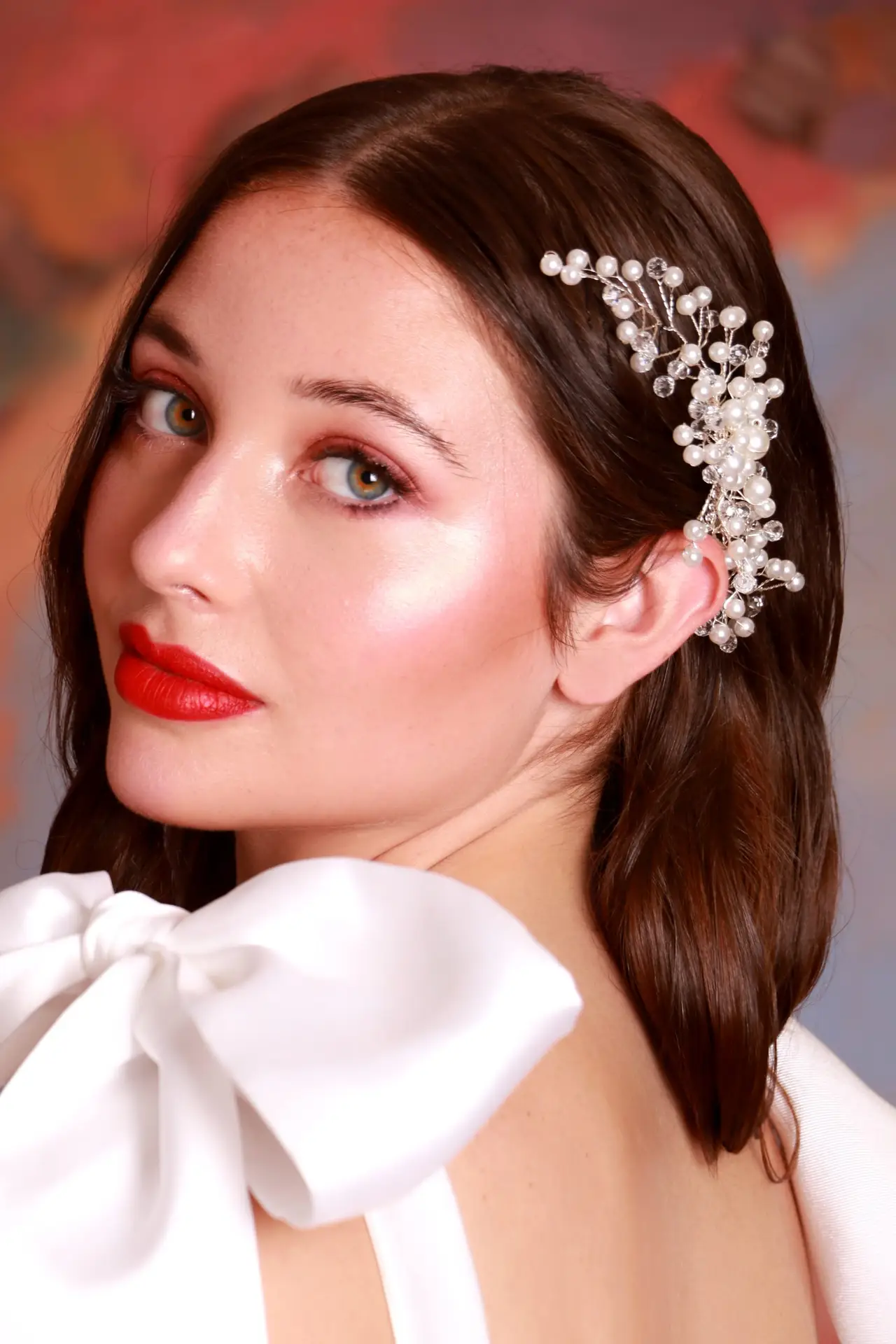 Portrait of a woman with elegant bridal makeup featuring bold red lips, styled brown hair with decorative pearl hair clip, wearing a white garment against a warm gradient background in Port Moody