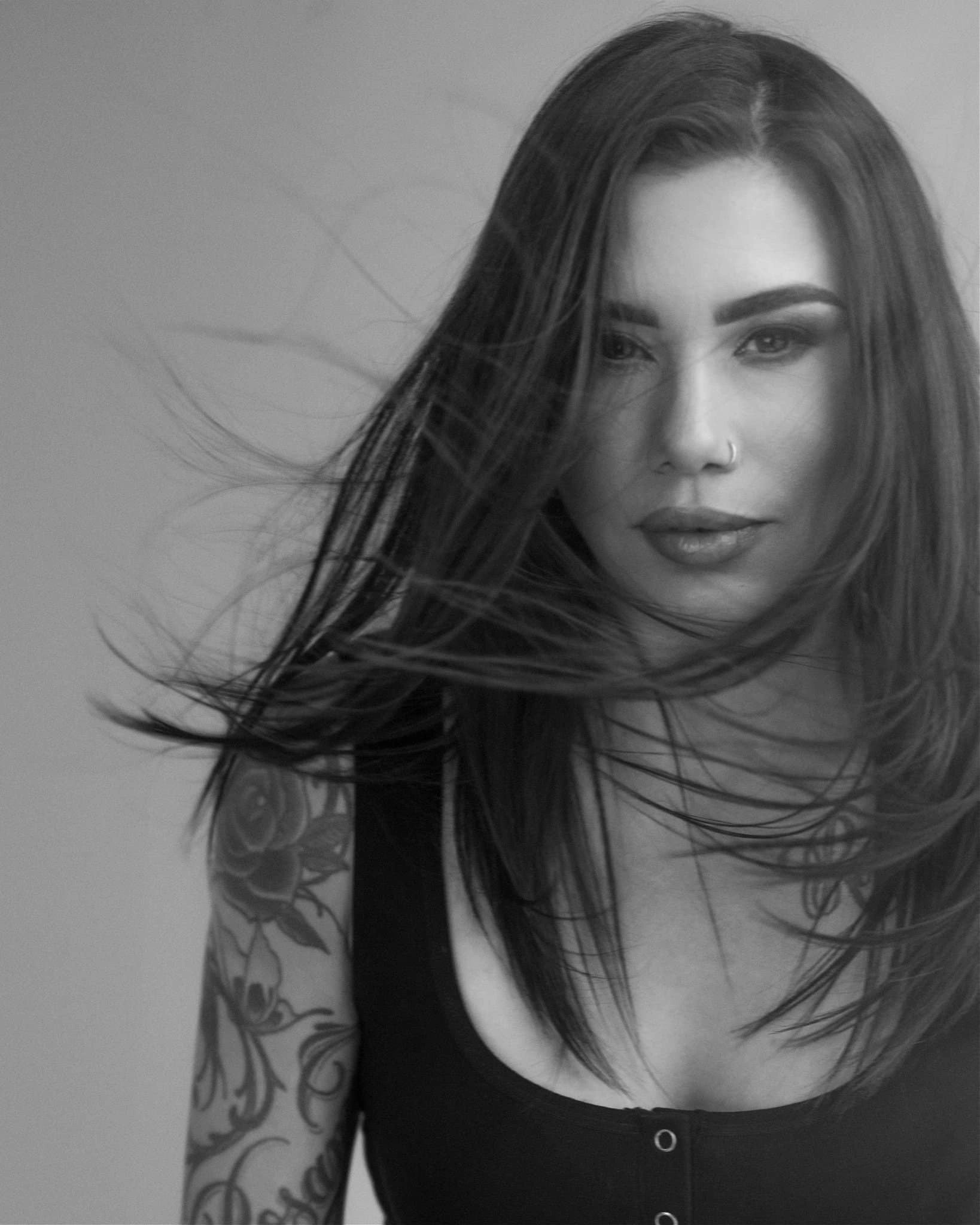 Woman with long dark hair and visible tattoos on her arm, shown in a black and white portrait photograph