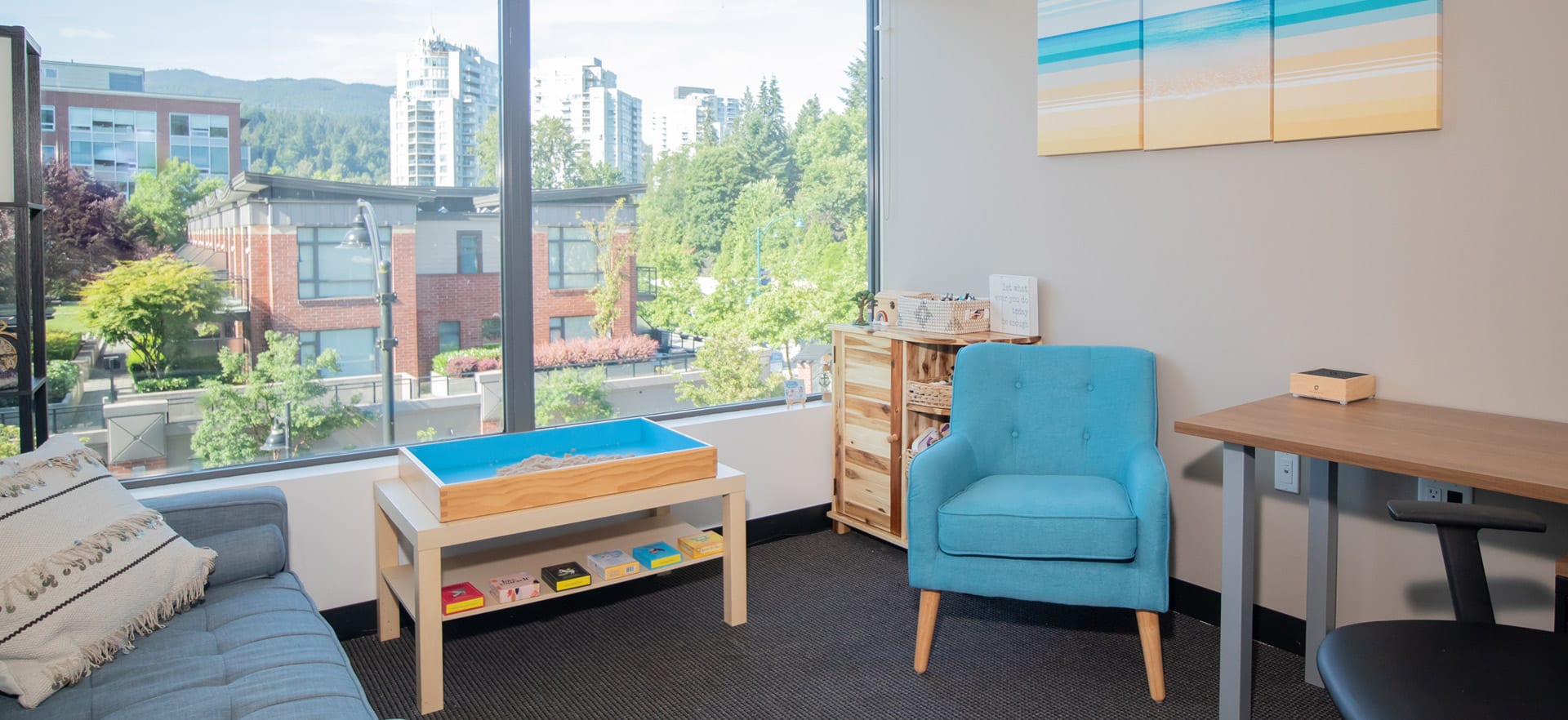 Modern pediatric counselling office in Port Moody with blue play table, turquoise armchair, natural wood furniture, and floor-to-ceiling windows overlooking the city and mountains