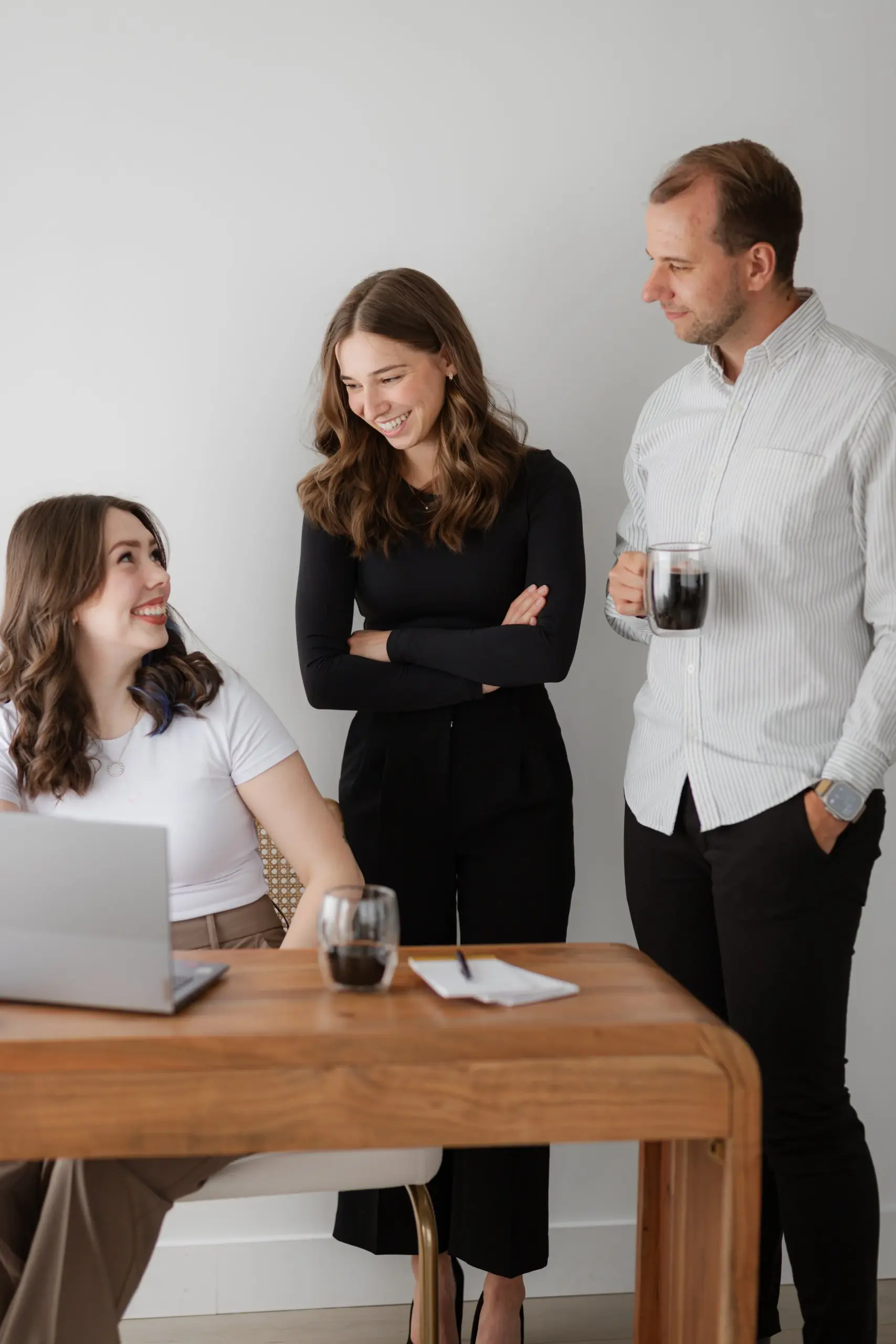 Three professionals in business attire engaged in conversation around a wooden table with a laptop and water glasses in a Port Moody office setting