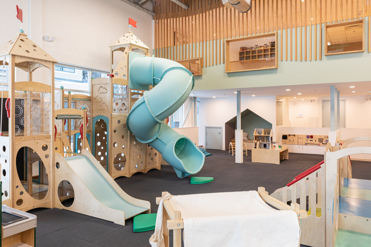 Modern indoor play space at Little Beans Play Cafe in Port Moody featuring a large blue spiral slide, wooden climbing structures with castles, and various play equipment on a dark floor with a mezzanine level