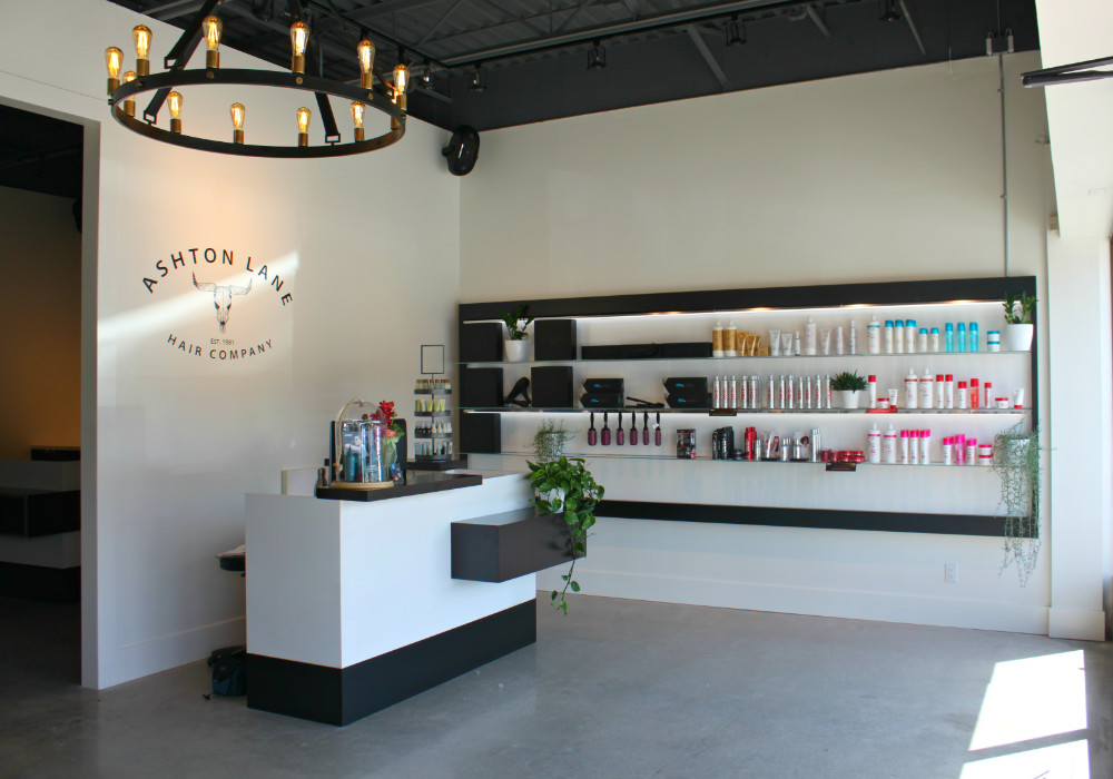 Ashton Lane Hair Company salon interior in Port Moody featuring a modern reception desk with black and white design, wall-mounted shelving displaying hair care products in various colors, and an elegant circular chandelier with warm lighting overhead
