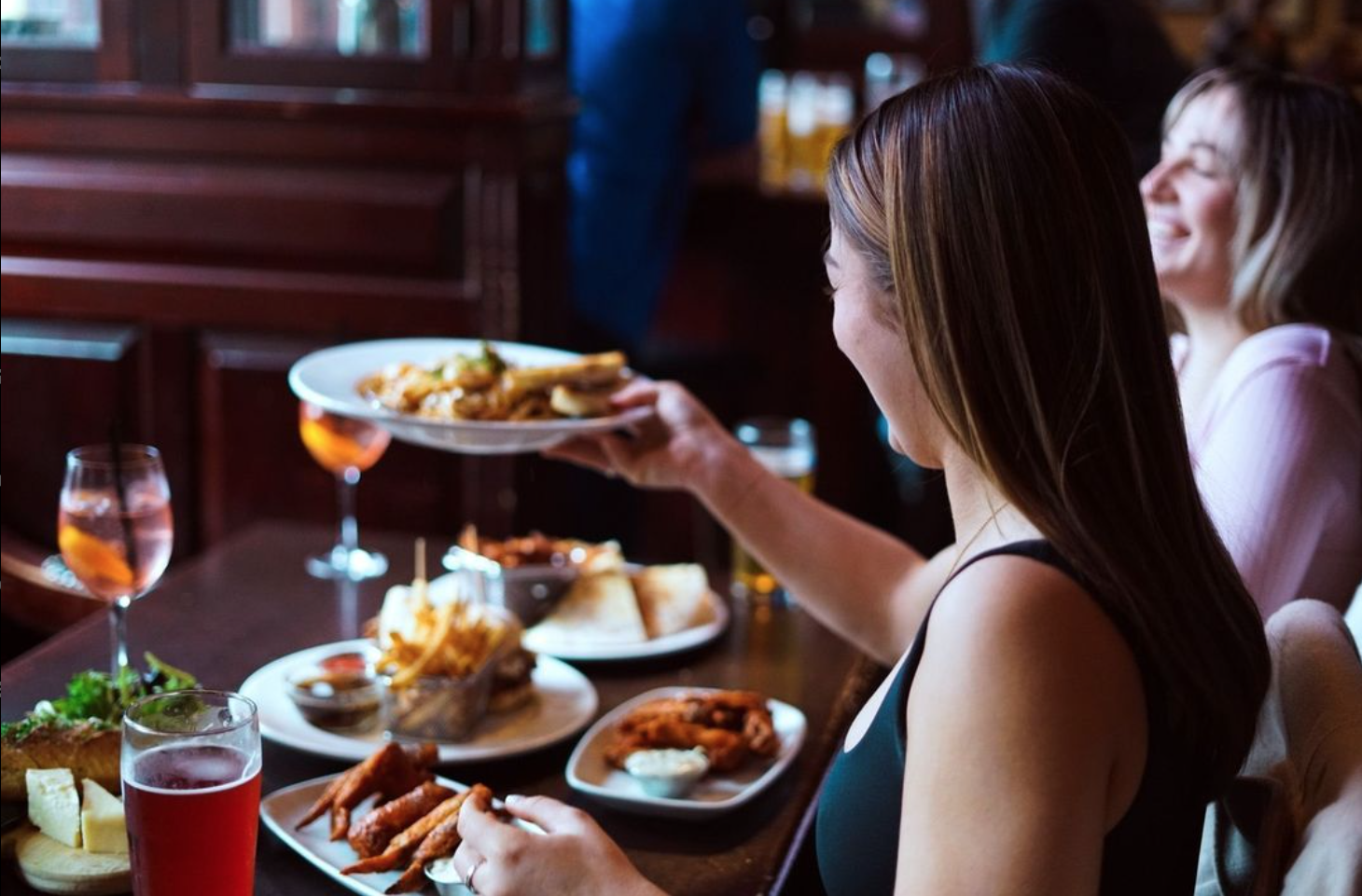Two women enjoying appetizers and drinks at a table in St James Well Pub in Port Moody, with plates of fried food, burgers with fries, and cocktails visible