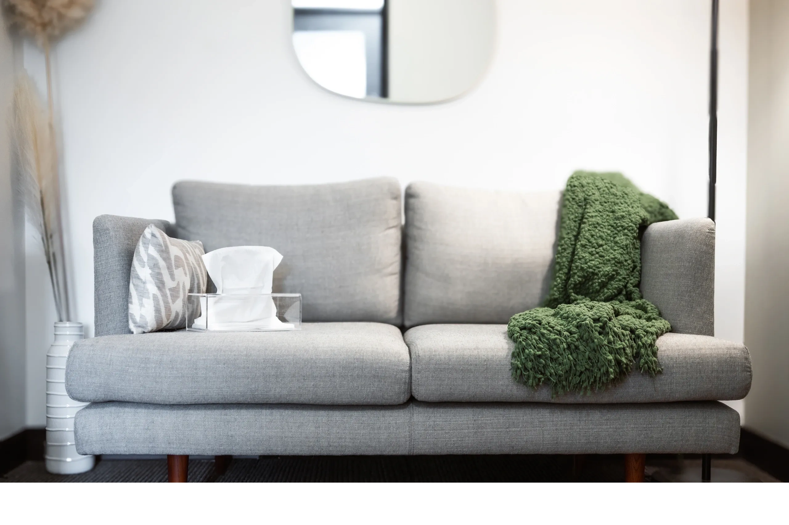 Modern minimalist living room with a gray upholstered sofa, green throw blanket, decorative pillows, white tissue box, and dried pampas grass in a vase, featuring a round mirror on the wall and bright natural lighting typical of contemporary Port Moody interior design