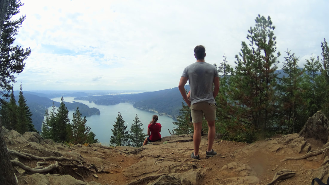 Two hikers standing on a mountain overlook with a scenic view of Burrard Inlet and Port Moody below, surrounded by evergreen trees