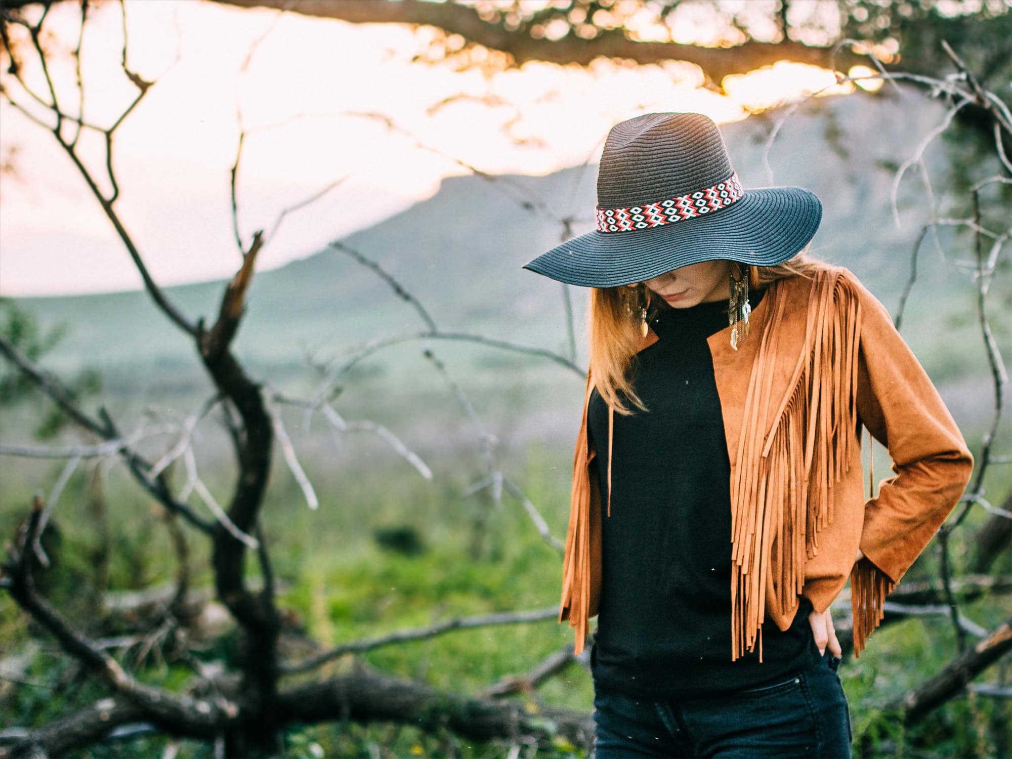 Woman wearing a blue wide-brimmed hat and tan fringe jacket standing in a mountainous landscape near Port Moody at golden hour