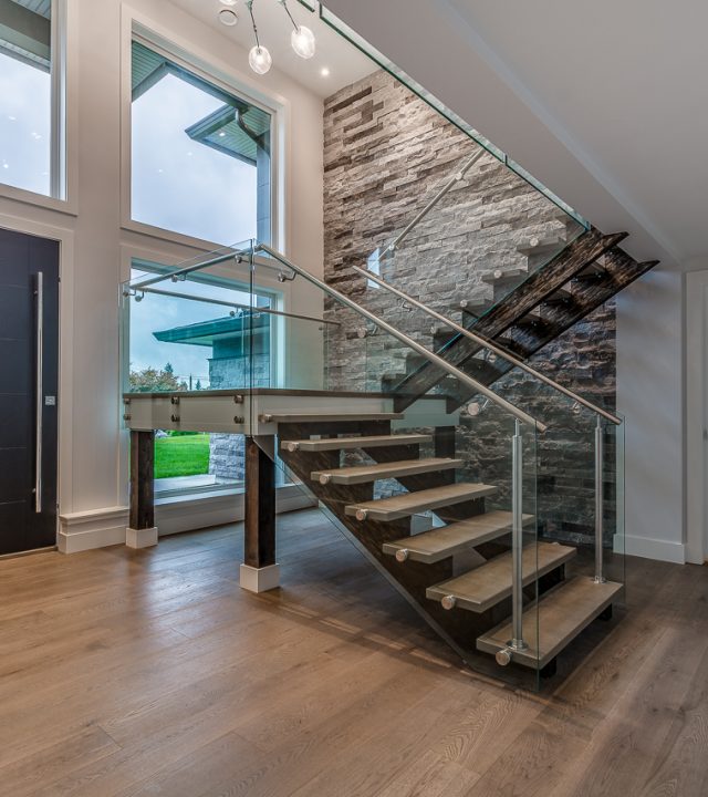 Modern staircase with glass railing and wooden steps in a Port Moody home renovation featuring stone accent wall and natural lighting