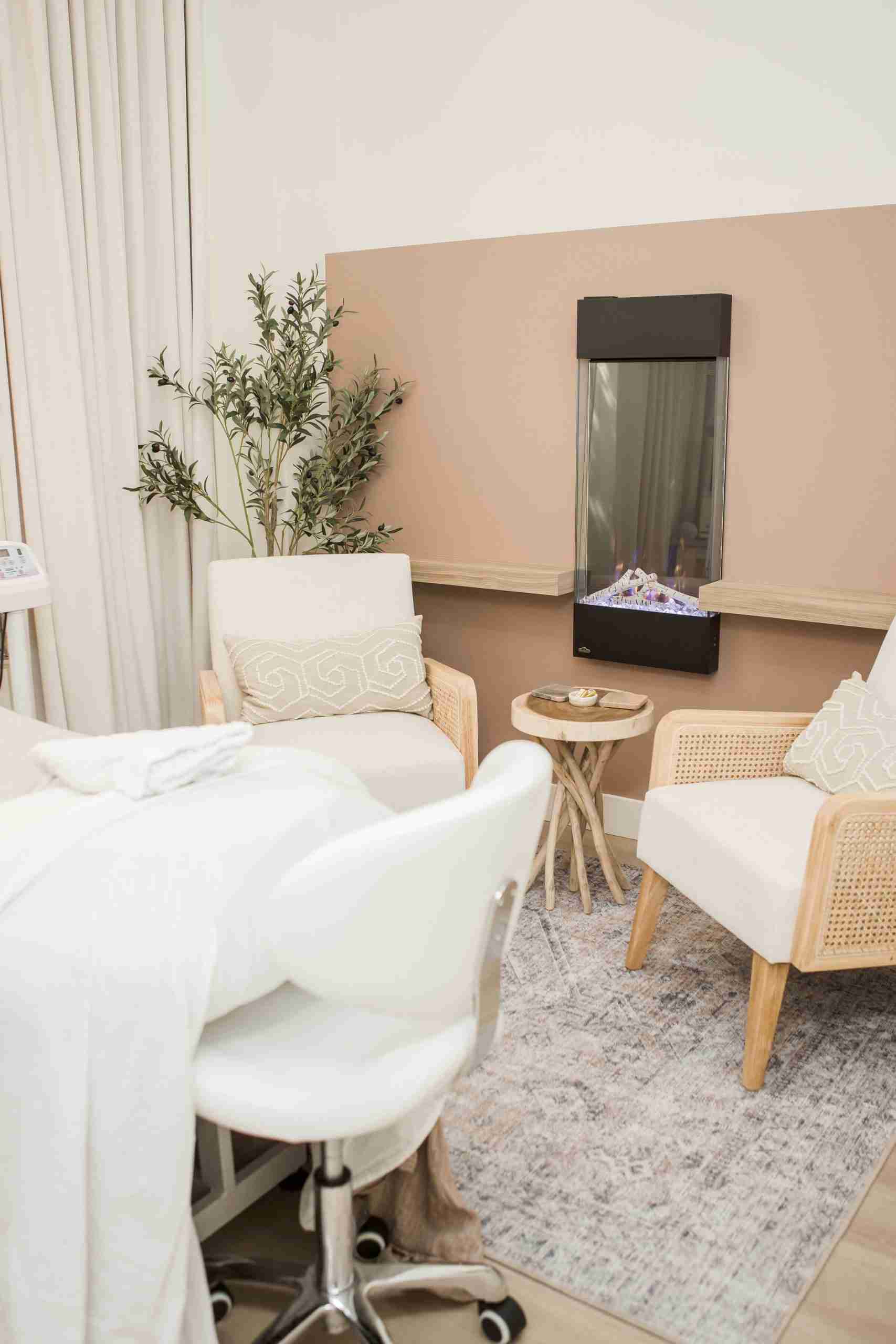 Modern somatic therapy room in Port Moody featuring two comfortable chairs with white cushions, a small round wooden side table, a potted plant, and a wall-mounted fireplace against a two-tone beige wall with floor-to-ceiling curtains