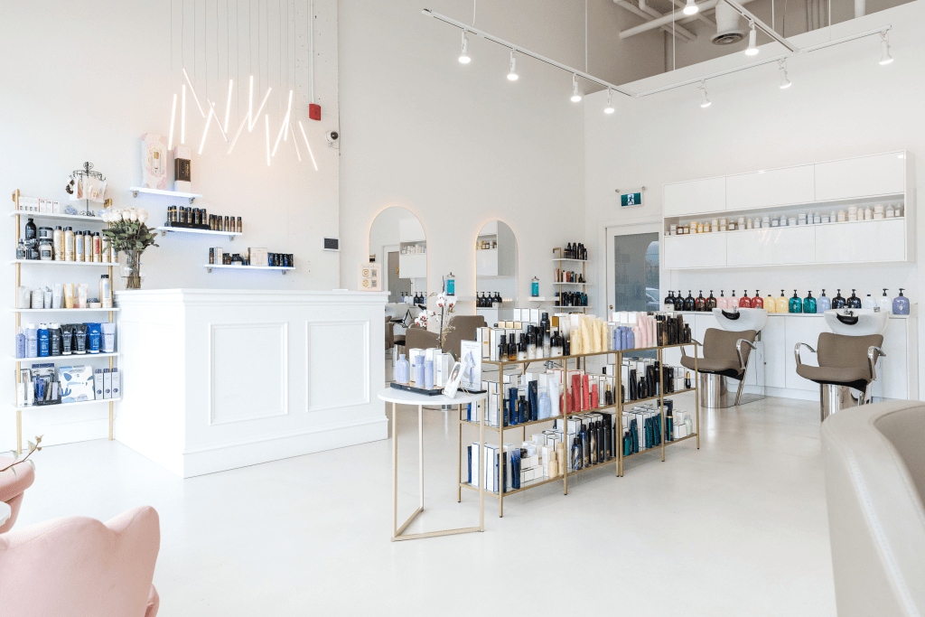 Modern, minimalist beauty salon interior in Port Moody featuring white walls, track lighting, display shelves with beauty products, a central product display table, salon chairs, and mirrors