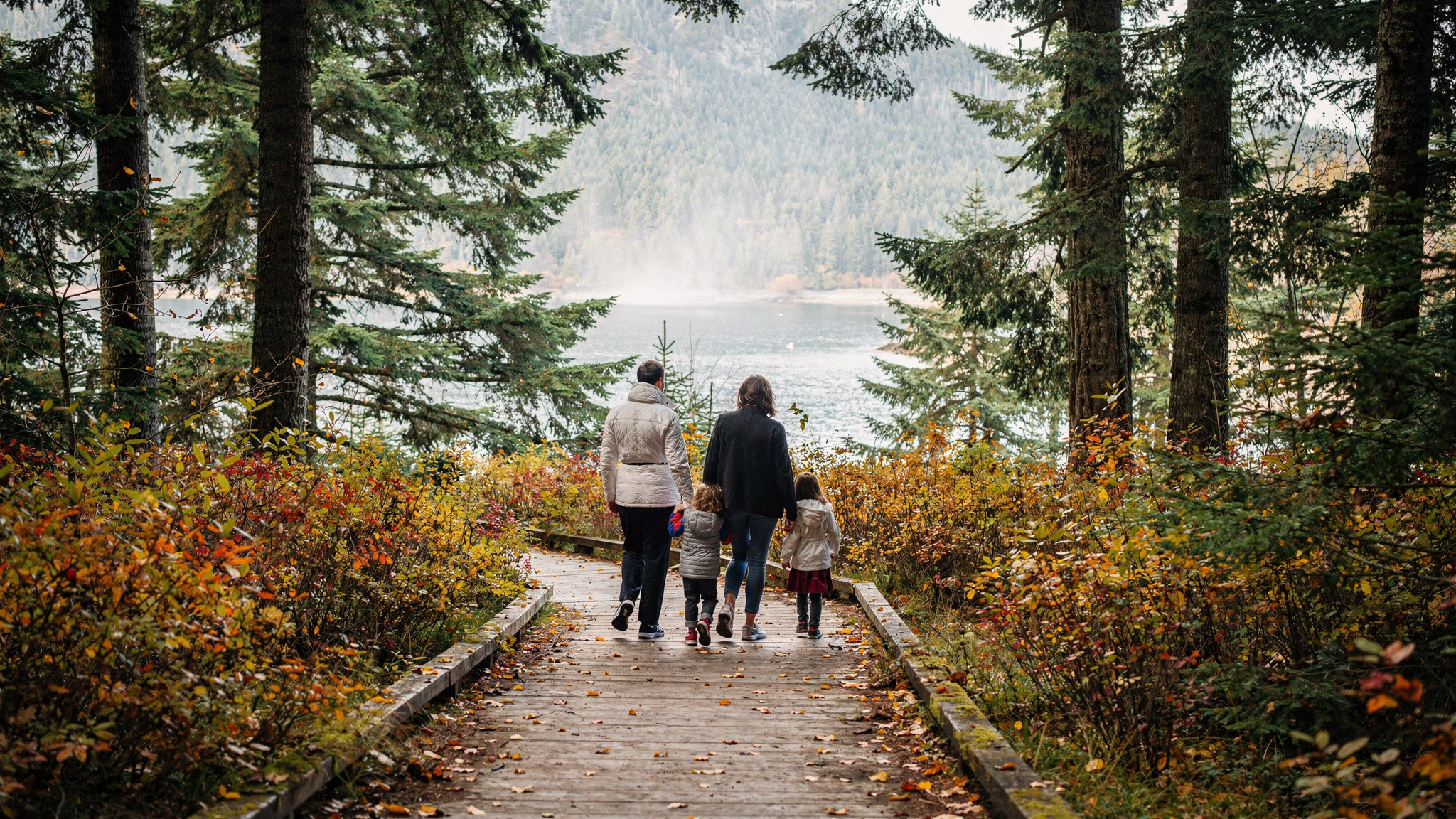 Family walking away from camera along a wooden boardwalk trail through forest near the water in Port Moody